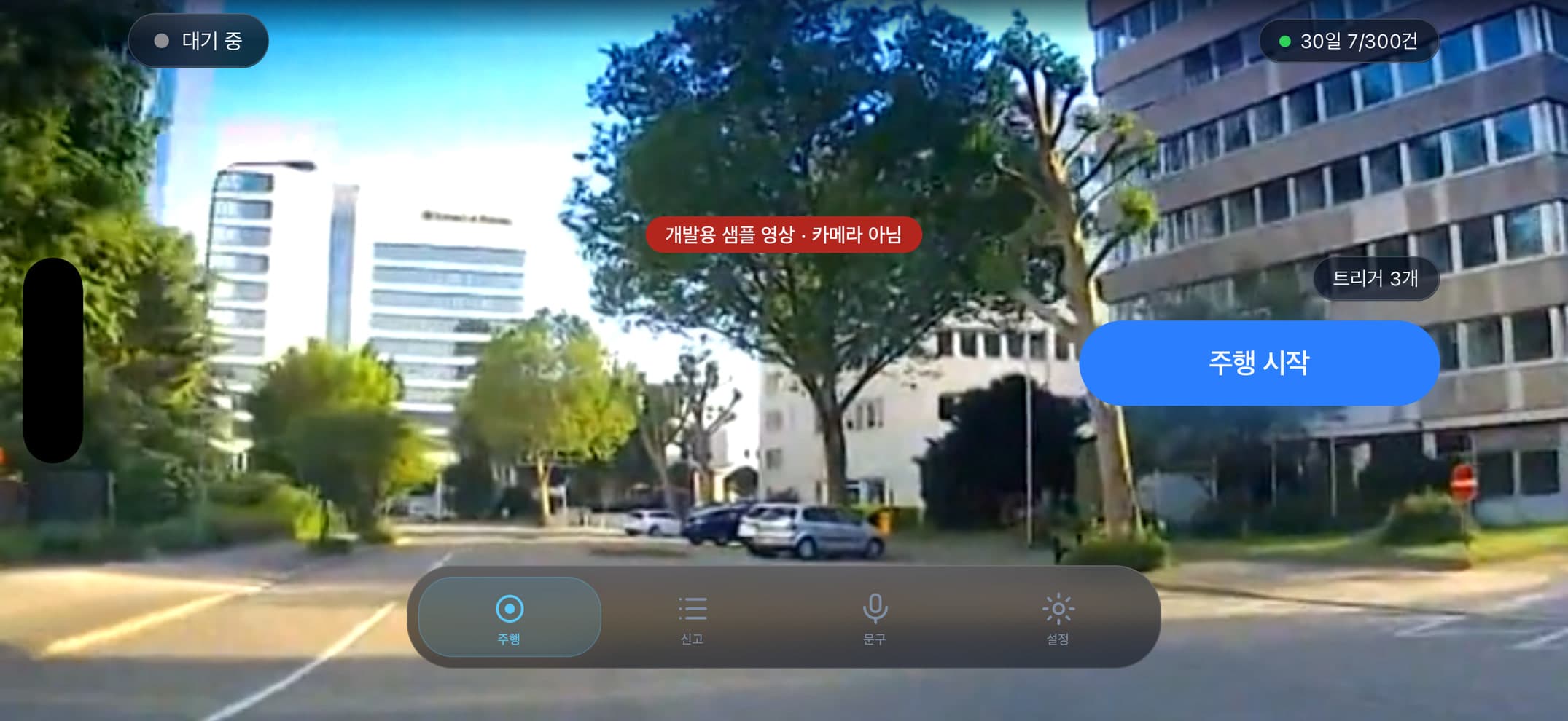 거치대에 가로로 올린 주행 화면 — 앞유리 너머 거리가 화면 전체를 채우고, 왼쪽 위에 "대기 중", 오른쪽 위에 30일 신고 한도, 오른쪽 가장자리에 "트리거 3개"와 파란 "주행 시작" 버튼. 영상 가운데에 "개발용 샘플 영상 · 카메라 아님" 배지.
